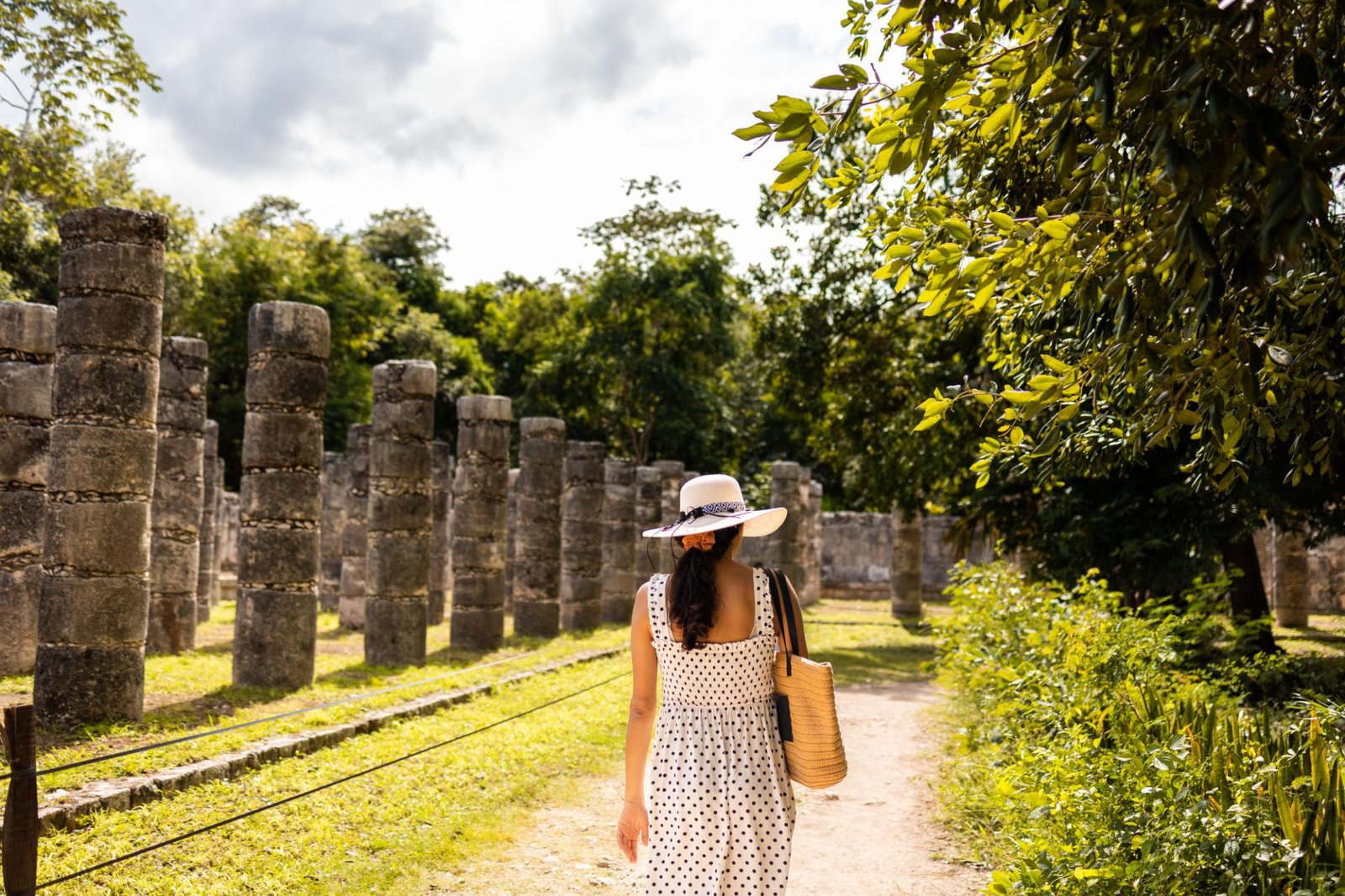Tourist woman in a white polka-dot dress and straw hat walks among ancient stone columns in a sunny outdoor ruin park.