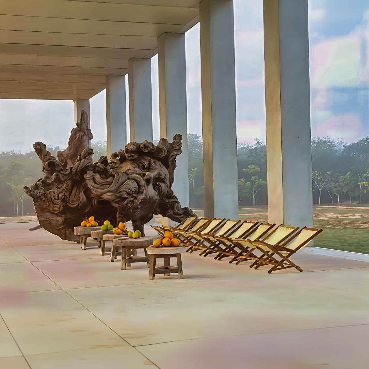 Yucatan-Art-Tours - authentictravelmexico.com Large abstract wooden sculpture on a covered patio with citrus fruit resting on small stools and a row of folding chairs nearby